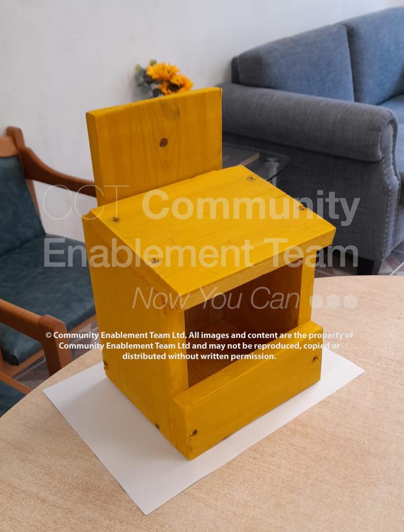 A small wooden birdbox painted in a bright yellow color sits on a table in a cozy living room setting. The birdbox has a simple design with an open front and a flat roof, showcasing visible screws at the corners. In the background, there's a grey sofa and a small bouquet of yellow flowers, adding a homely touch to the scene.
