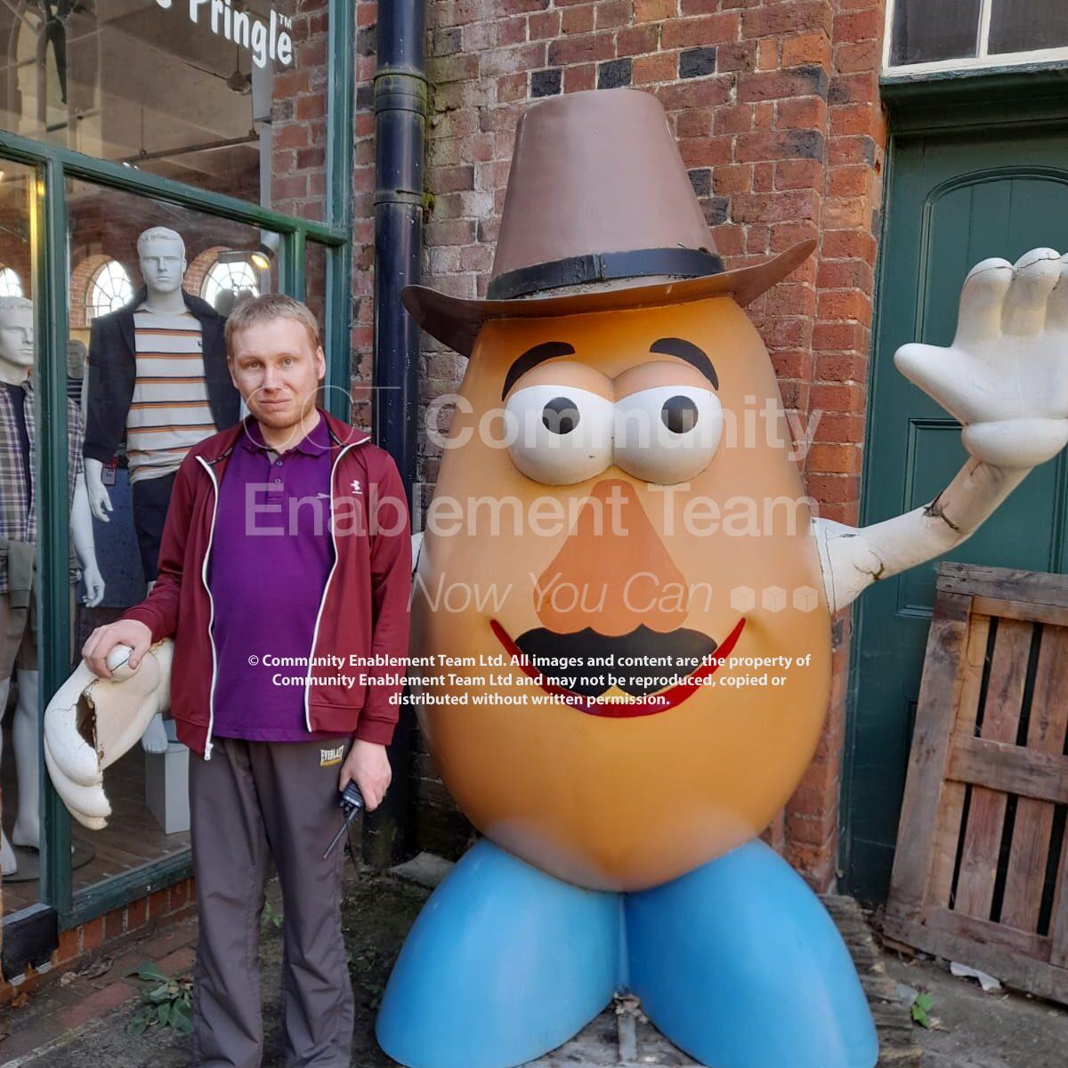 A man in a burgundy jacket and purple polo shirt standing next to a large, smiling potato statue wearing a cowboy hat, outside a James Pringle store.