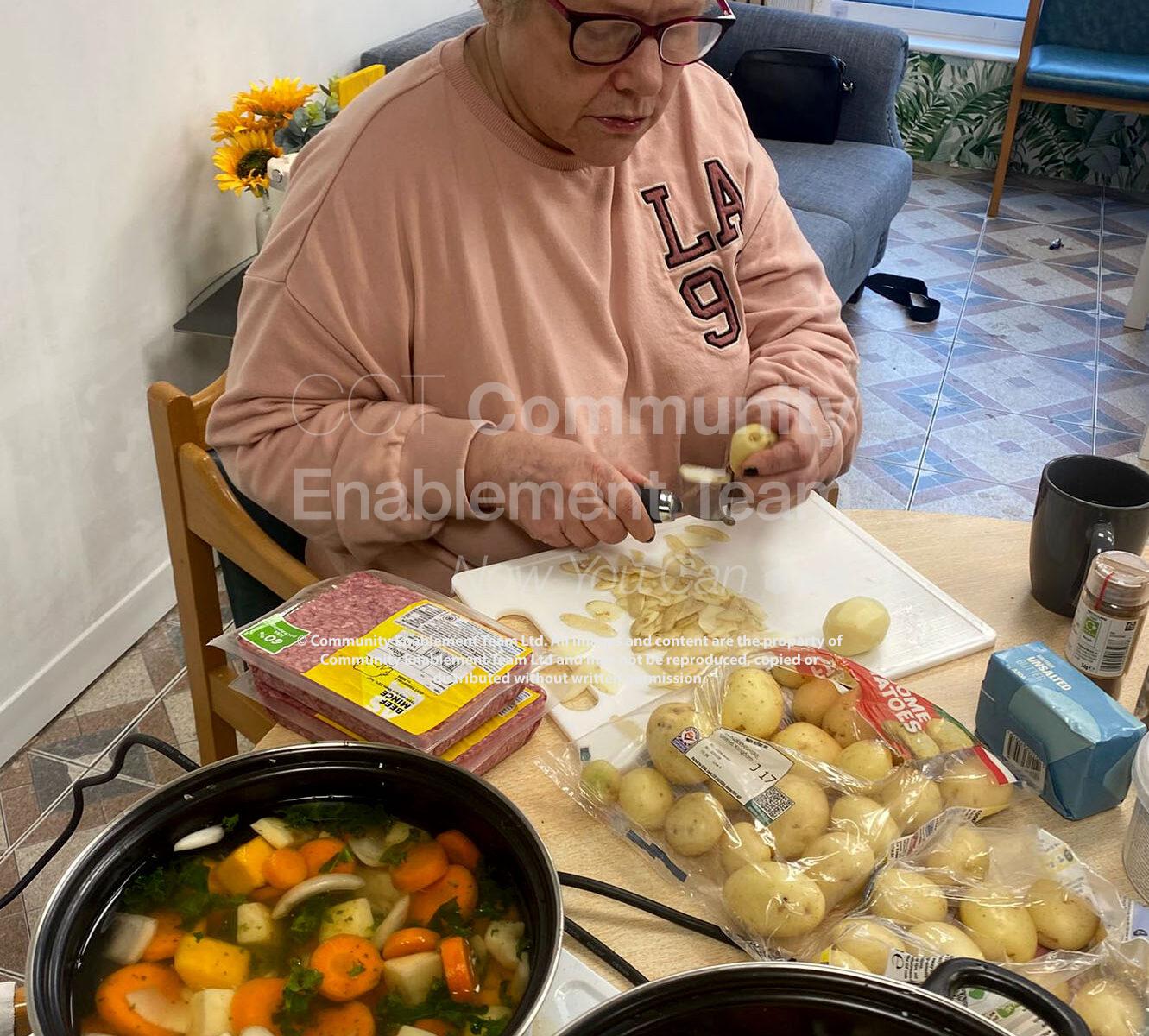 An older woman peeling potatoes at a table, surrounded by ingredients including carrots and potatoes. Two large pots of vegetable soup or stew are cooking on the table in front of her.