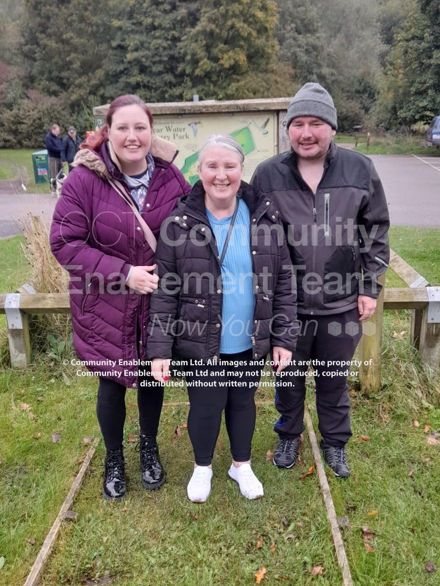 Three people standing together, smiling, in front of the Vicar Water Country Park sign. The woman in the center wears a black puffer jacket, while the other woman is in a purple coat, and the man is in a black jacket and grey beanie. The background shows the park’s green landscape.