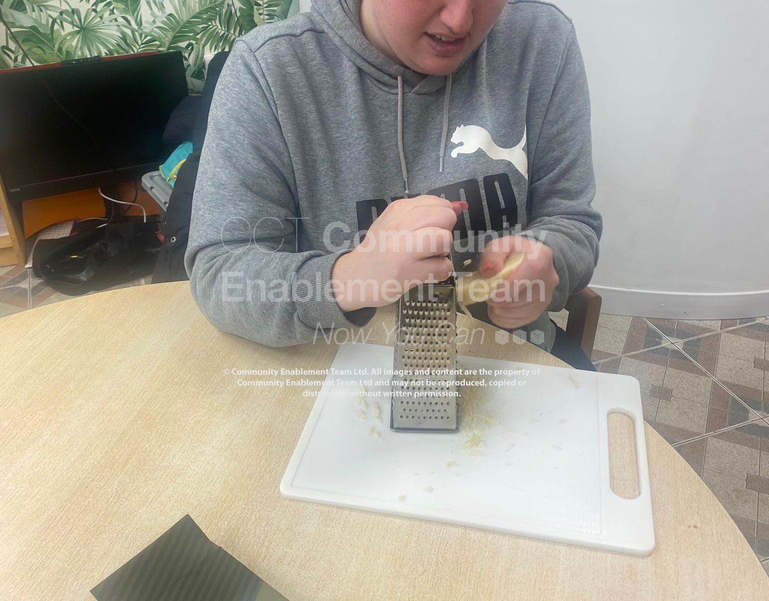 A woman in a grey Puma sweatshirt grates cheese on a white cutting board in preparation for cooking. A box of lasagne sheets and a baking tray sit on the table beside her.