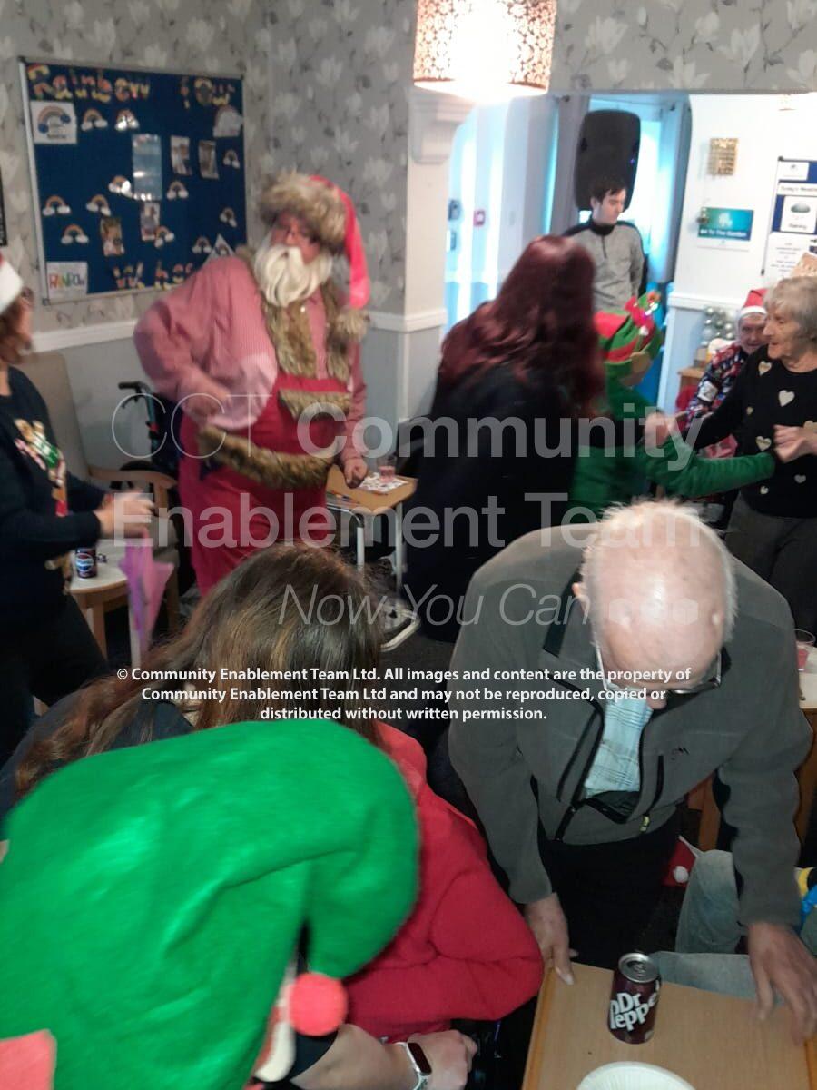 Santa Claus interacting with attendees at a lively Christmas celebration, with festive decorations, including a bulletin board and holiday posters, in the background.