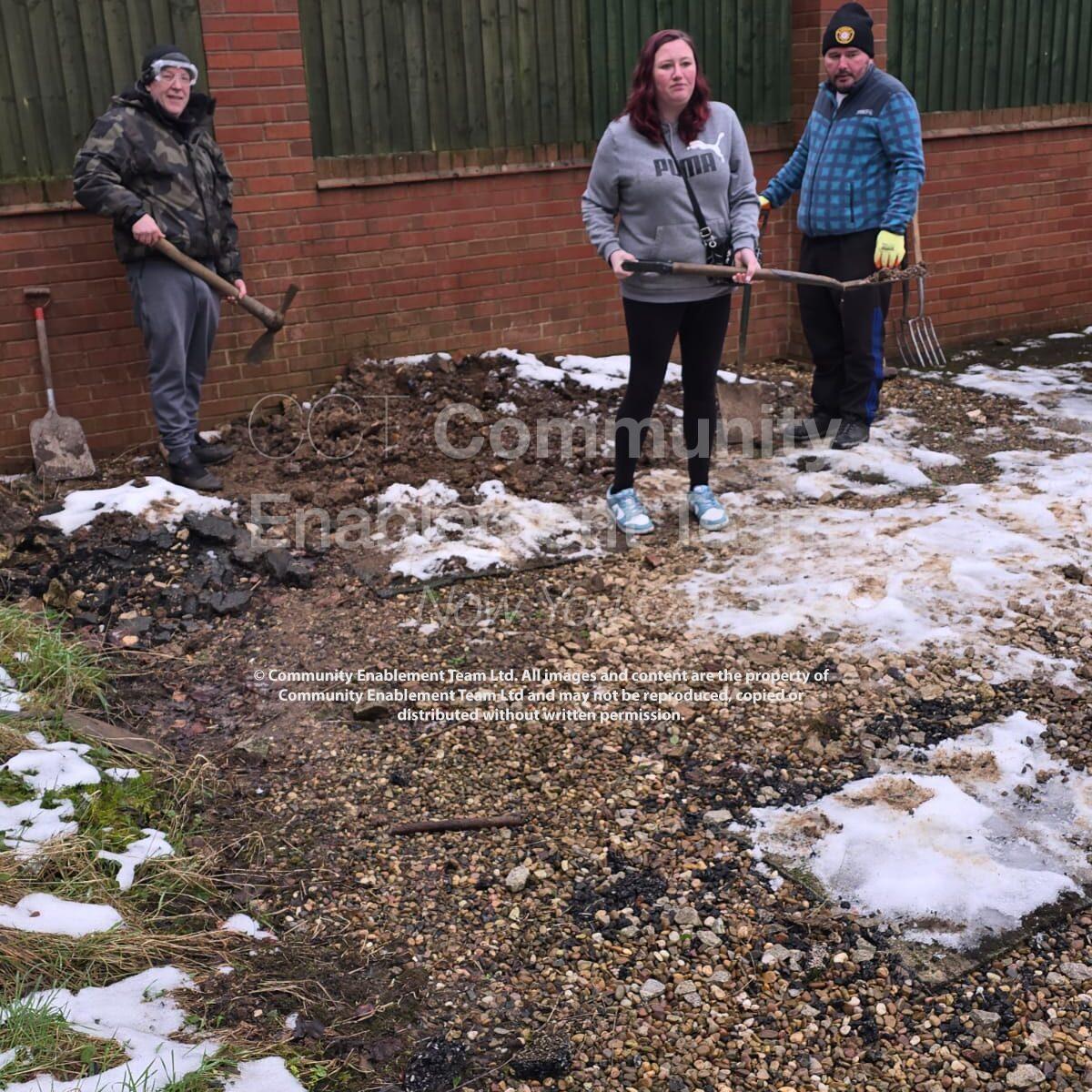 Three individuals working outdoors in a garden area during winter. The ground is covered with patches of snow, gravel, and dirt. One person is holding a pickaxe, another is holding a spade, and the third is holding a garden fork. A brick wall and wooden fence form the background.