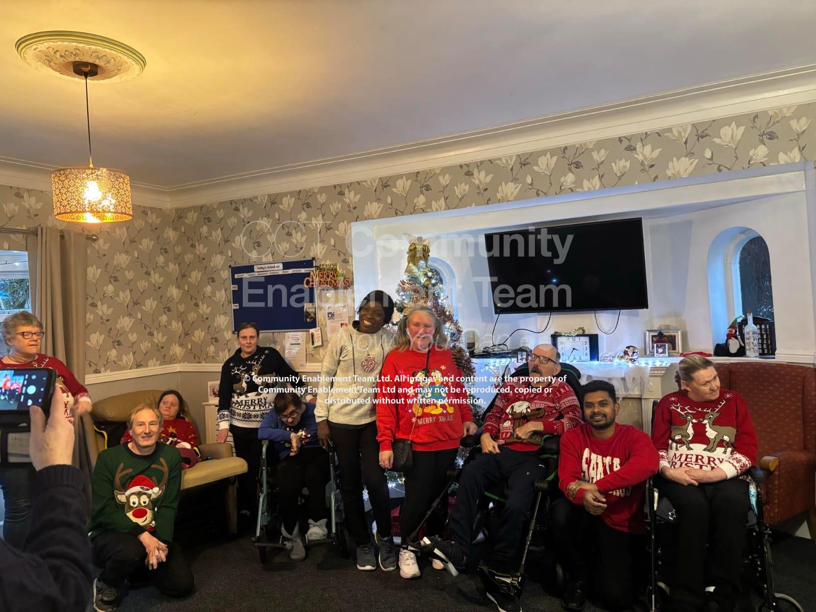 A group of residents and staff from the Community Enablement Team and Enablement Care posing together in festive Christmas jumpers. The group is gathered in a warmly decorated room with a Christmas tree and holiday decor in the background, radiating holiday cheer.