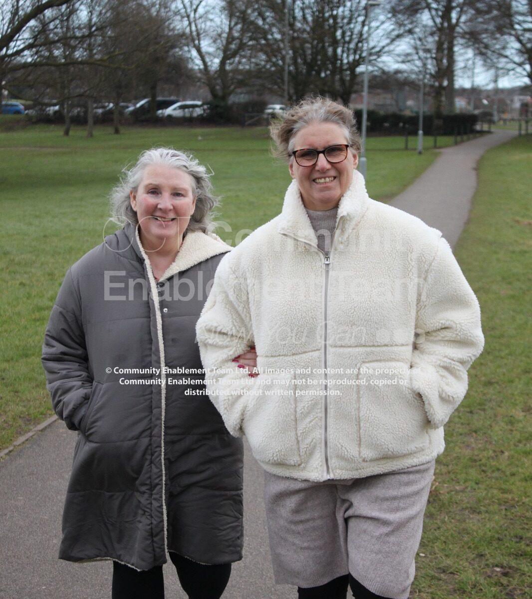 The same two women are walking arm in arm through an open park. They smile while strolling along a wide pathway lined with grass, trees, and distant houses.