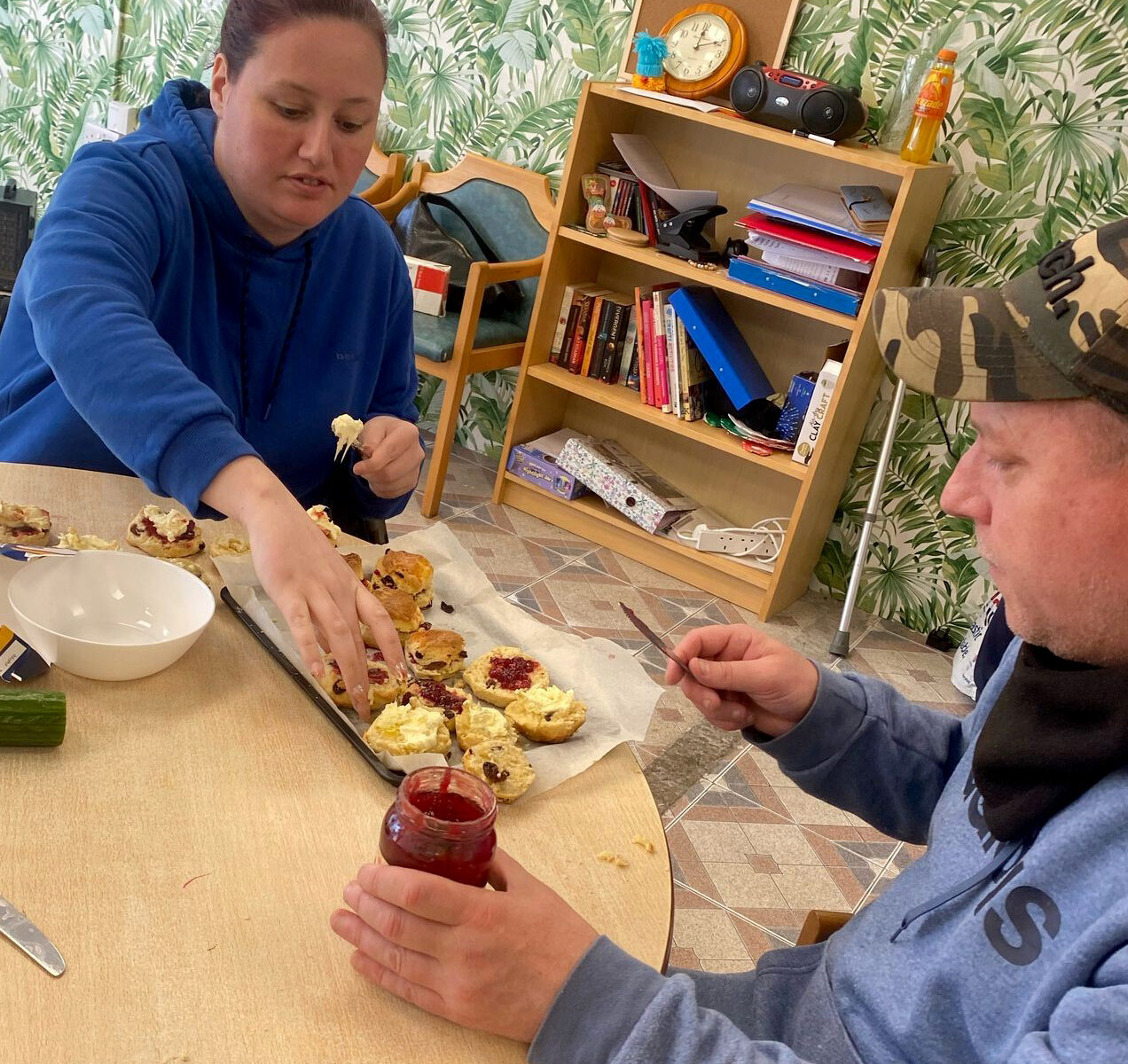A man and a woman sat at a table preparing scones with cream and jam