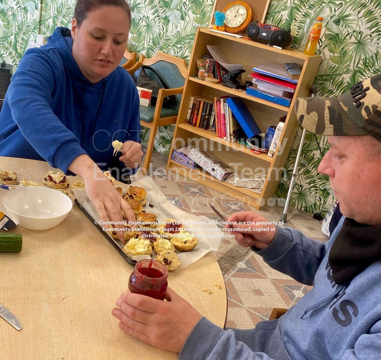 A man and a woman sat at a table preparing scones with cream and jam