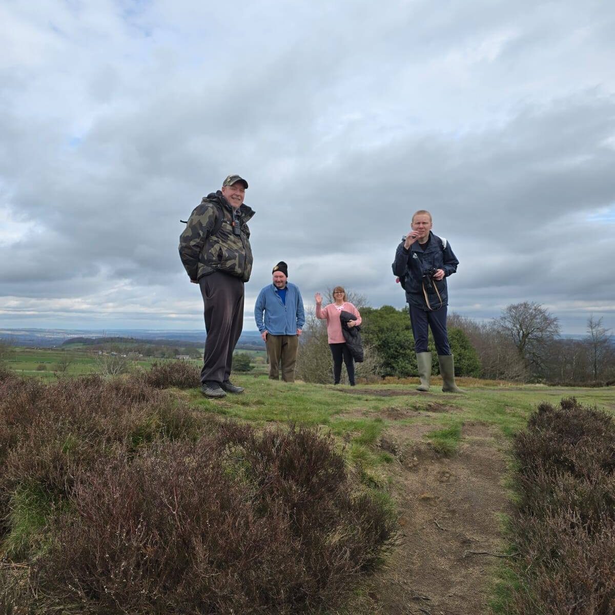 Four walkers stand smiling on a grassy hilltop surrounded by heather under a cloudy sky. The group appears cheerful and relaxed, with expansive countryside views stretching out behind them.