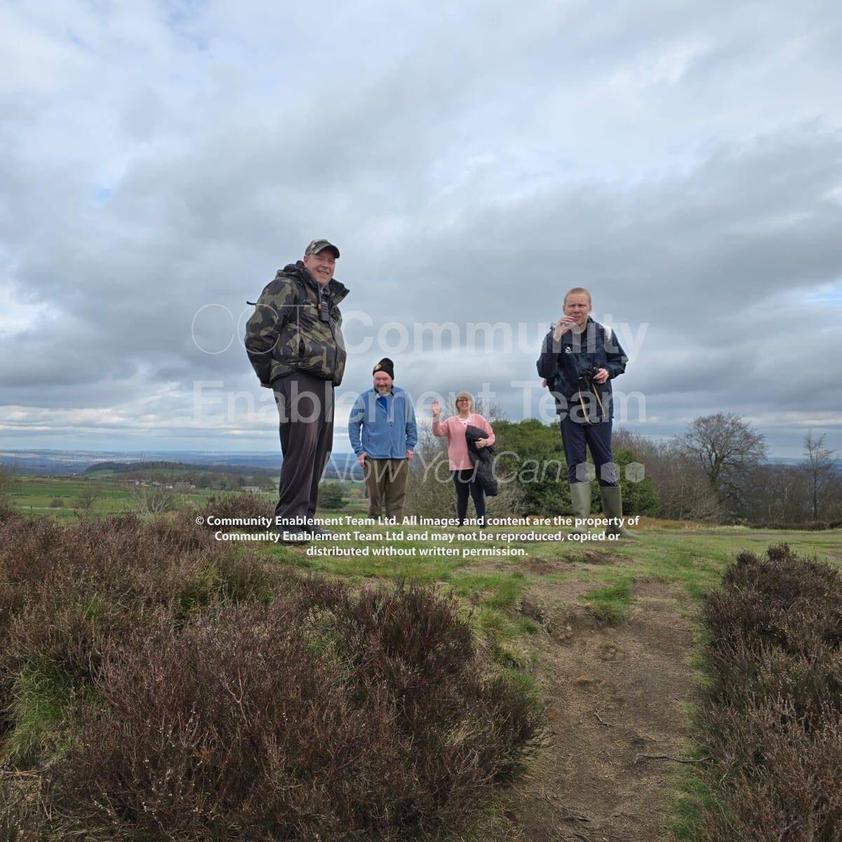 Four walkers stand smiling on a grassy hilltop surrounded by heather under a cloudy sky. The group appears cheerful and relaxed, with expansive countryside views stretching out behind them.