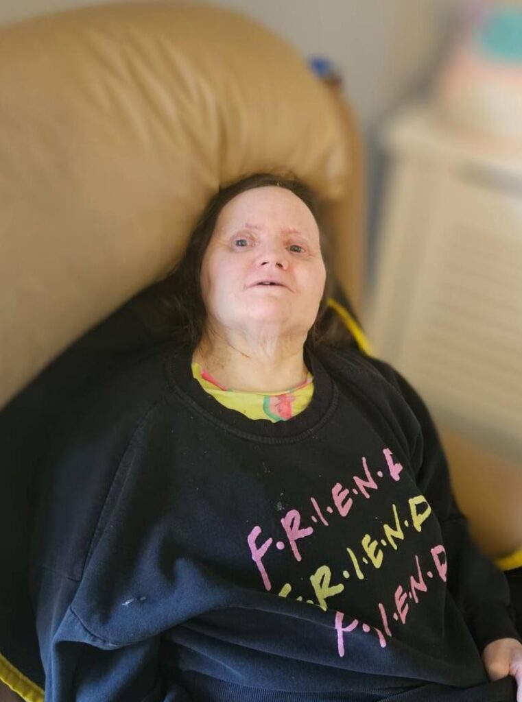 A woman seated comfortably in an armchair, smiling and wearing a sweatshirt with the word "FRIENDS" repeated in colourful text.