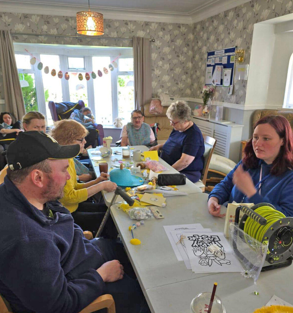 Participants seated at a long table in a cosy room, focused on crafting Easter bonnets and cards. Decorative bunting and soft lighting add to the warm atmosphere.