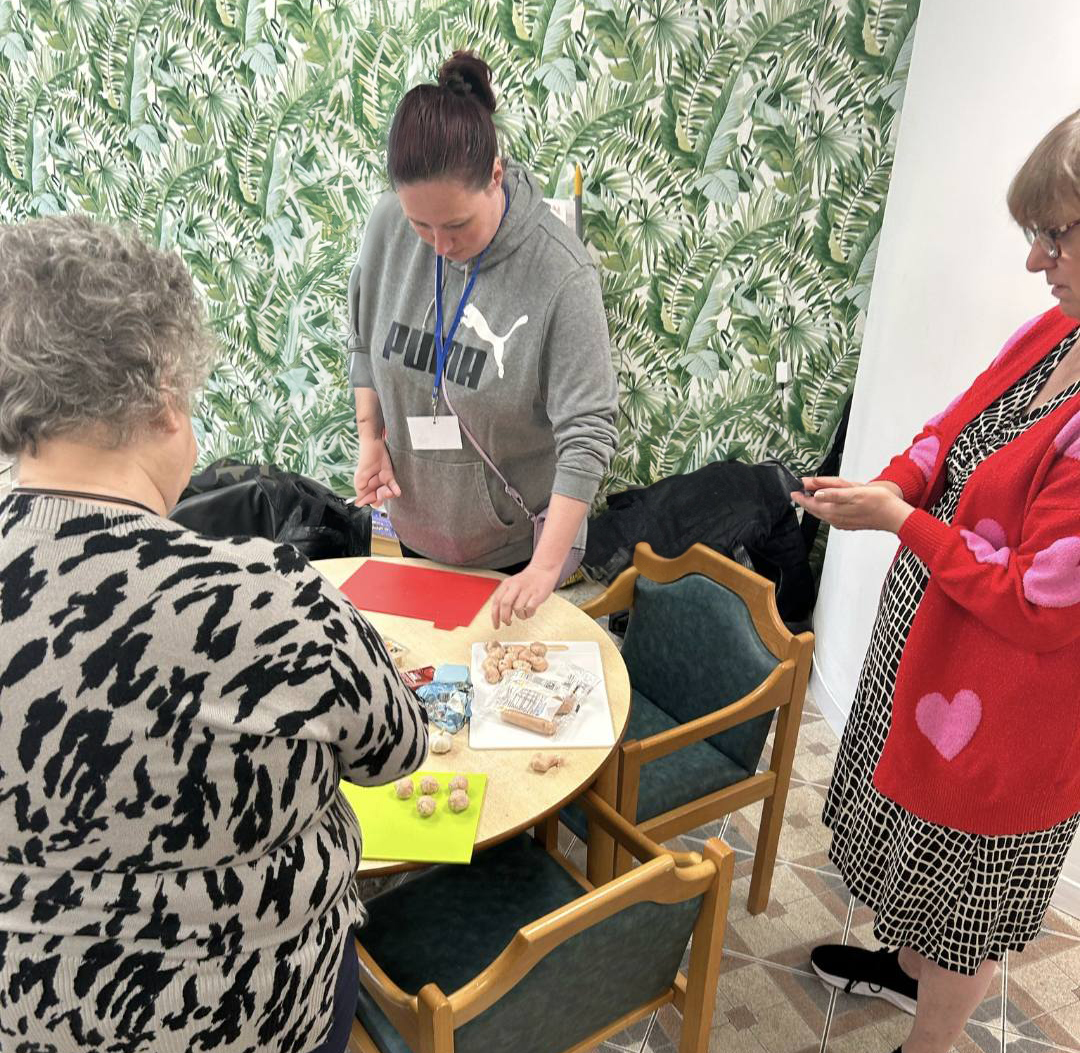 Three women gather around a table preparing ingredients, including meatballs and packaging. One woman chops on a red board while the others assist, set against a green leafy wallpaper background.