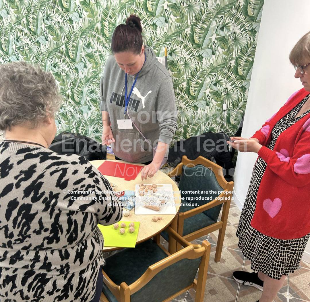 Three women gather around a table preparing ingredients, including meatballs and packaging. One woman chops on a red board while the others assist, set against a green leafy wallpaper background.
