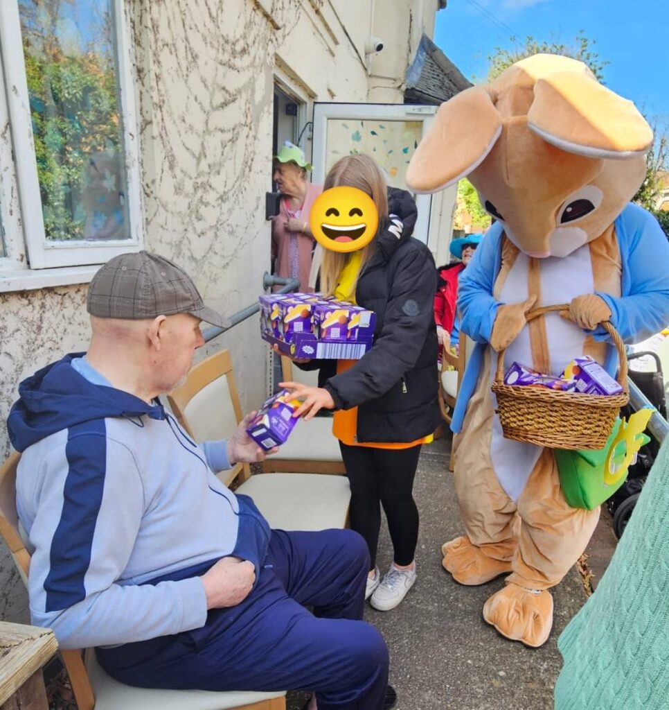 A person in an Easter Bunny costume handing out chocolate eggs to residents seated outside, assisted by a smiling volunteer.