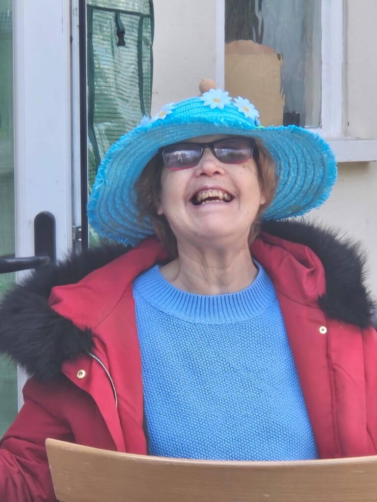 A woman wearing a bright blue hat decorated with daisies, smiling widely in a red coat and blue jumper.