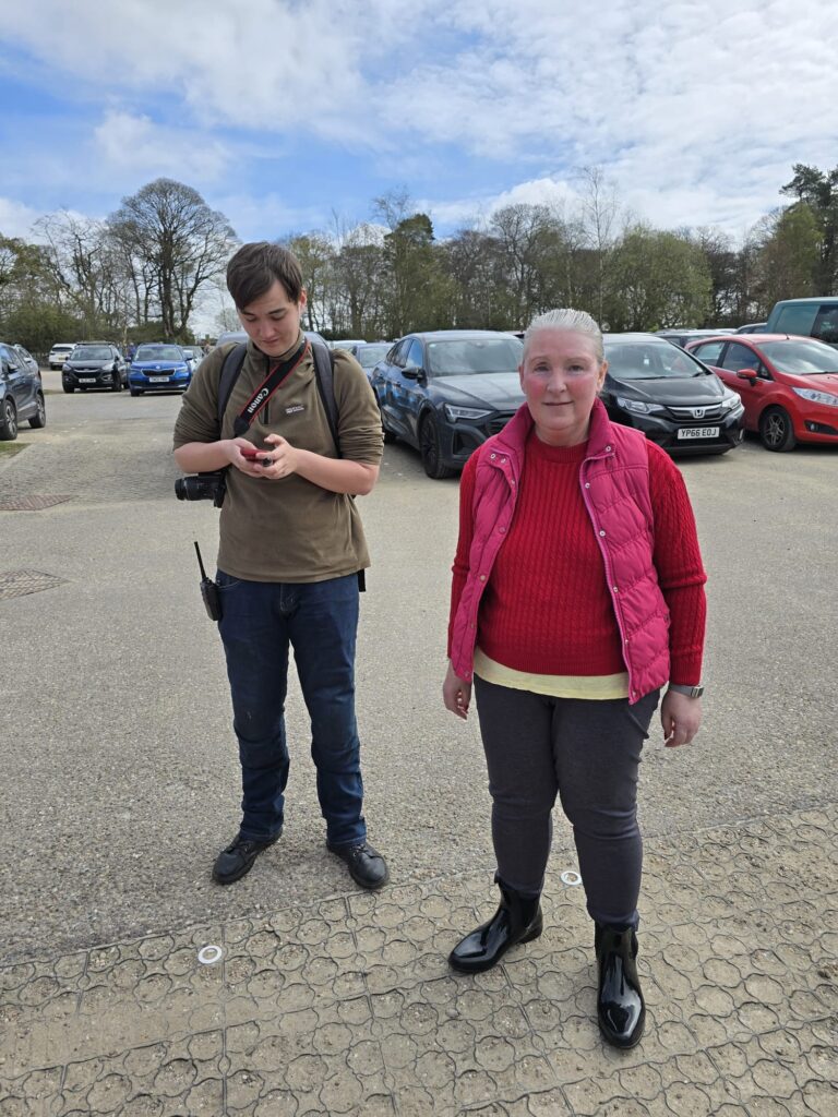 Two participants in the car park after the walk – one checking a phone with a camera around his neck, the other smiling toward the camera.
