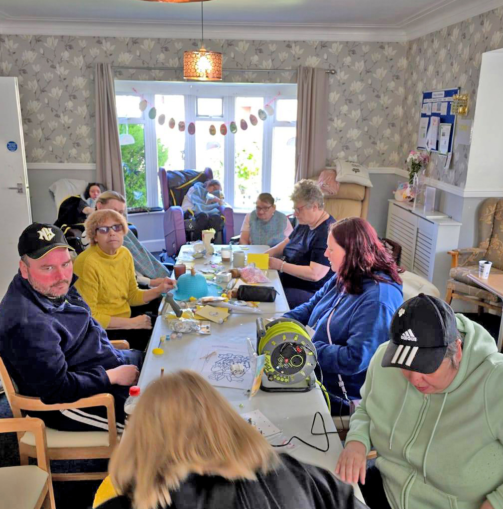A wide shot of the full group working on crafts together at a long table. The room is decorated with Easter-themed bunting and natural light pours in from the windows.
