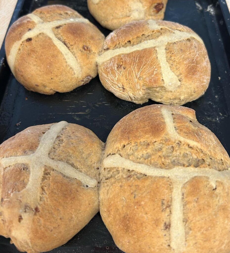A close-up of freshly baked hot cross buns on a tray, golden brown with visible crosses.