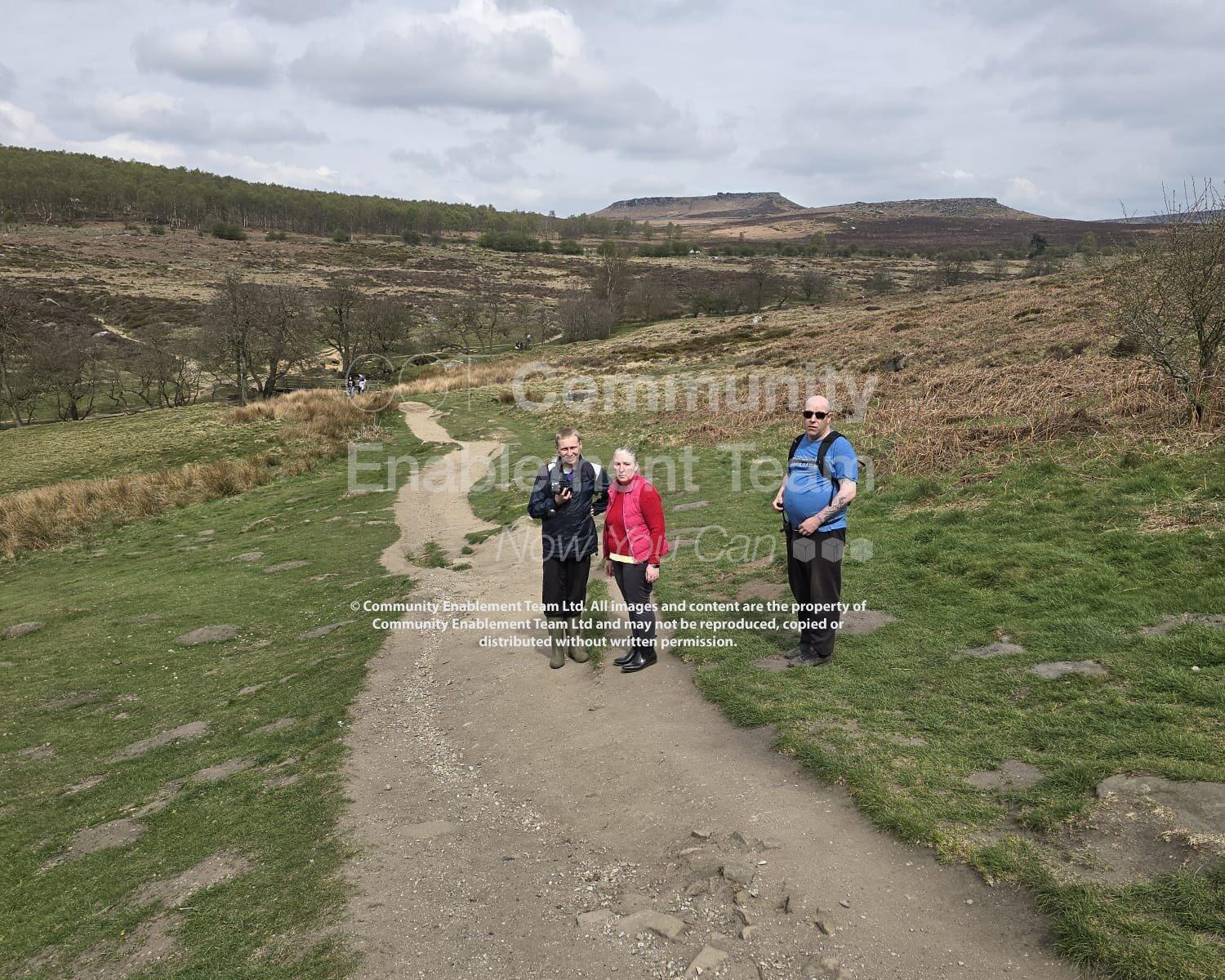 Three individuals from CET’s Walking Club standing on a wide footpath at Padley Gorge, surrounded by rolling moorland and hills under a lightly clouded sky.