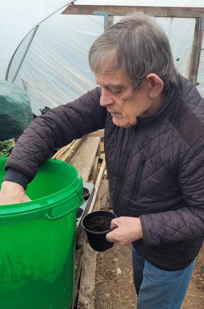 Man potting compost into plant pots – A man fills black plastic pots with compost from a green bucket inside a polytunnel.