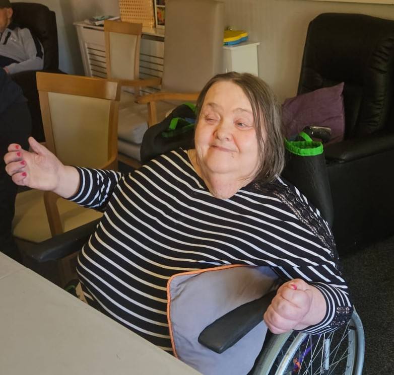 A woman in a wheelchair indoors, smiling warmly while seated at a table.