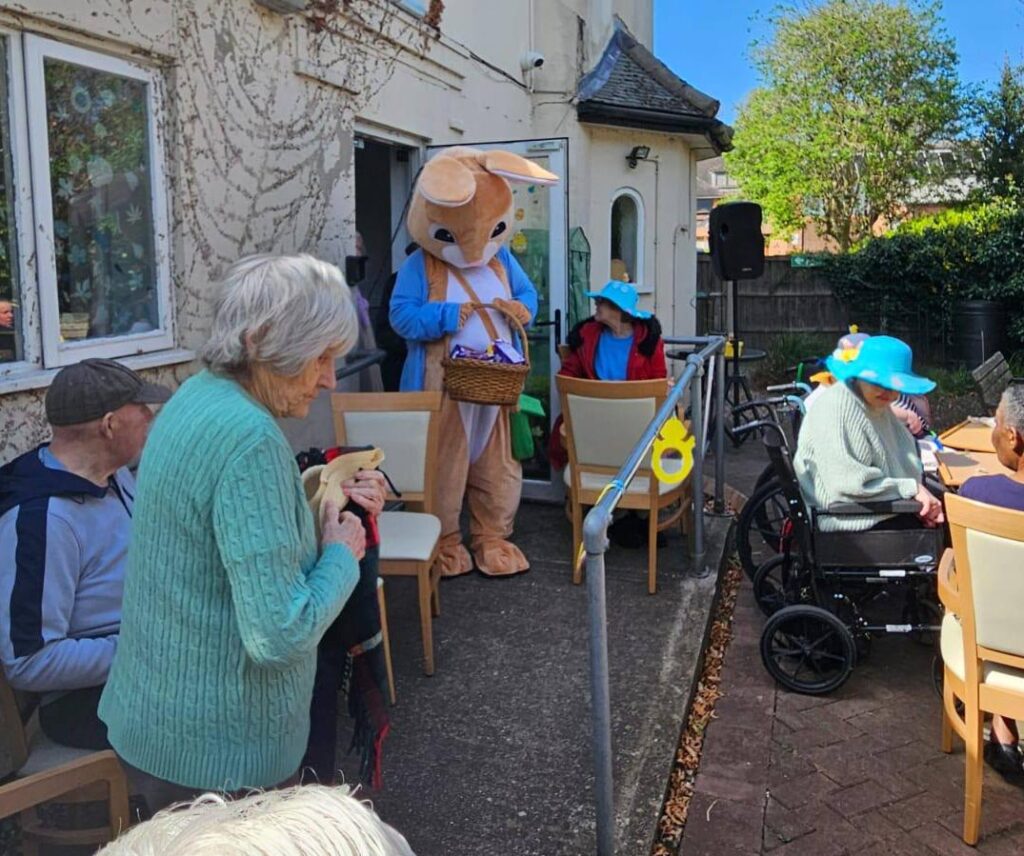 The Easter Bunny standing at the doorway with a basket of eggs, as residents sit nearby ready to take part.

