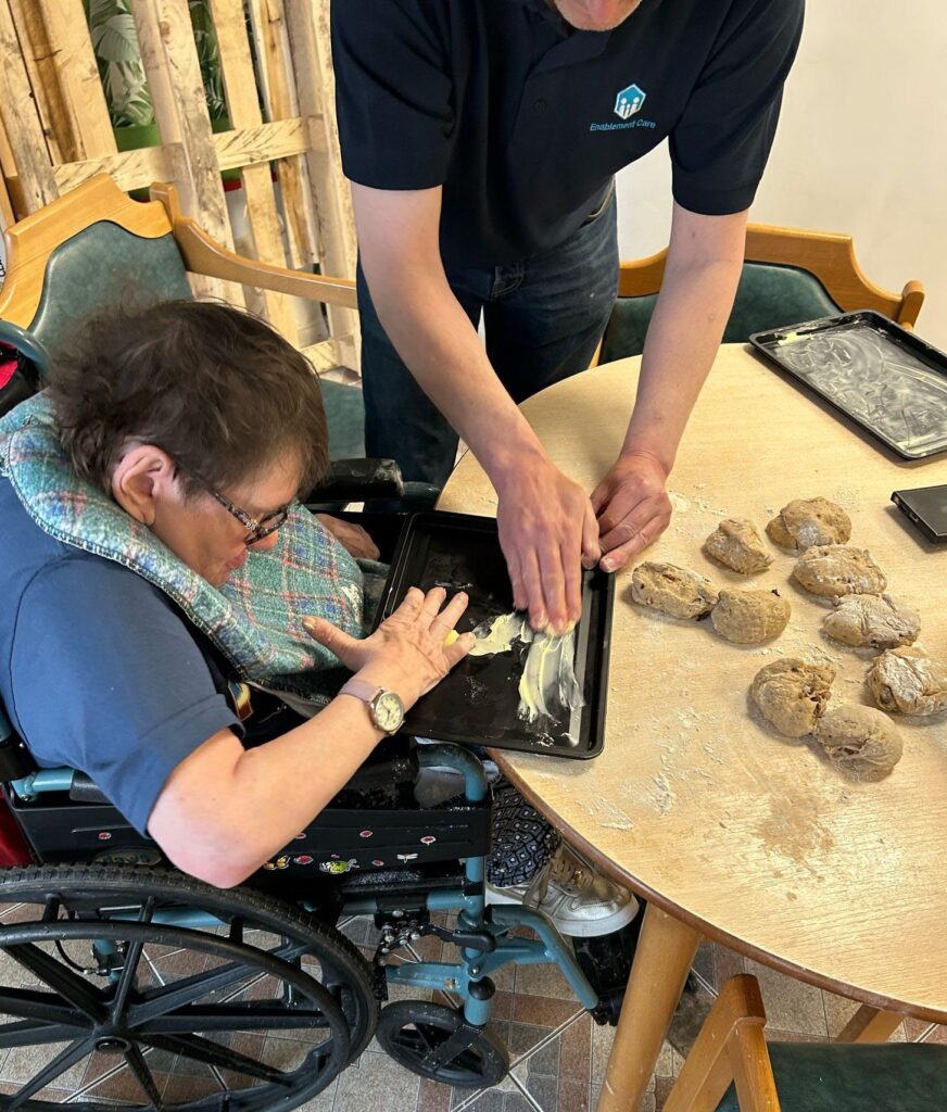 A woman in a wheelchair and a support worker greasing a baking tray together, with prepared dough balls on the table.