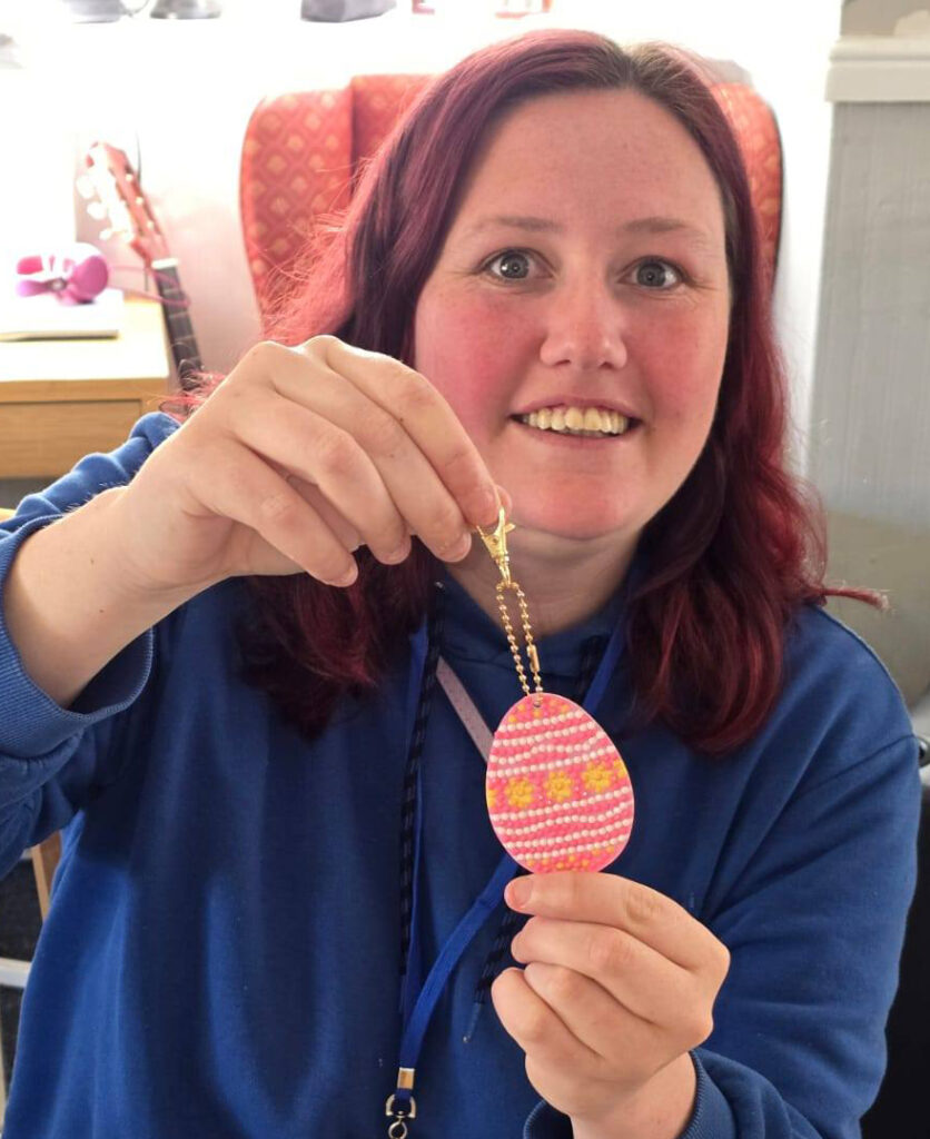 A smiling woman holding up a beaded Easter egg keyring she has just finished crafting.