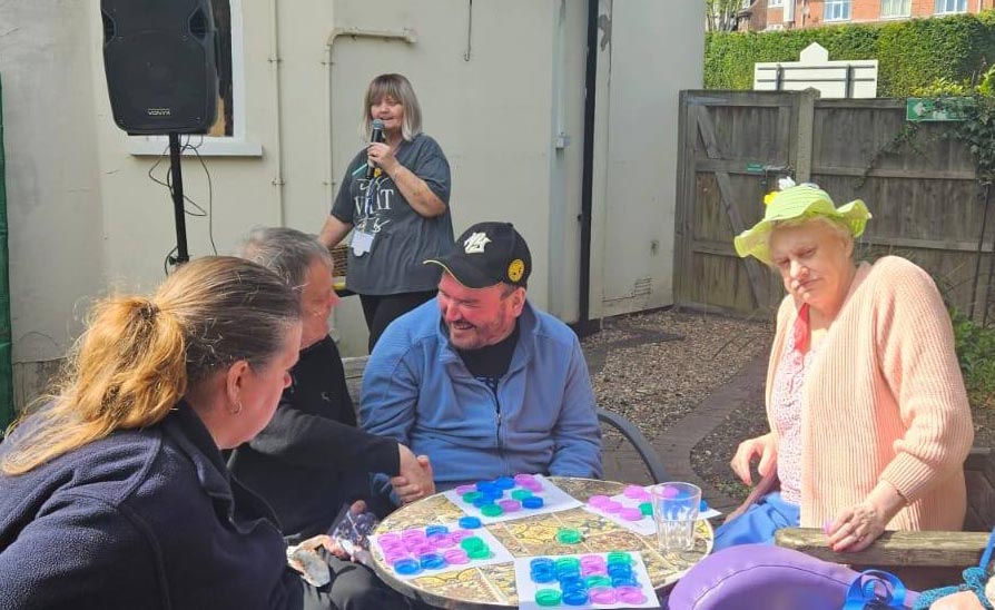 A group of residents playing bingo around a table outside, smiling and chatting, with a staff member speaking into a microphone in the background.