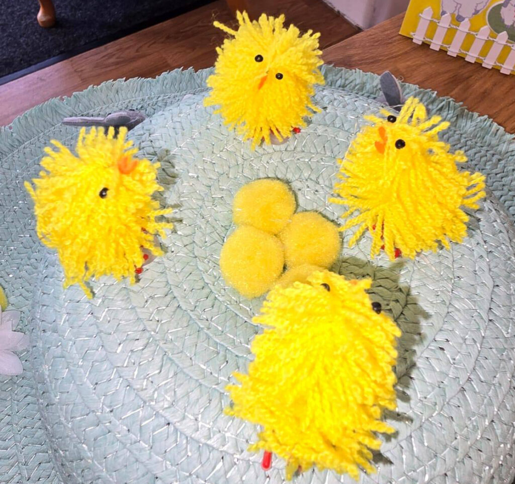 Close-up of a pastel green Easter bonnet decorated with bright yellow pom-pom chicks and additional pom-poms arranged in the centre.