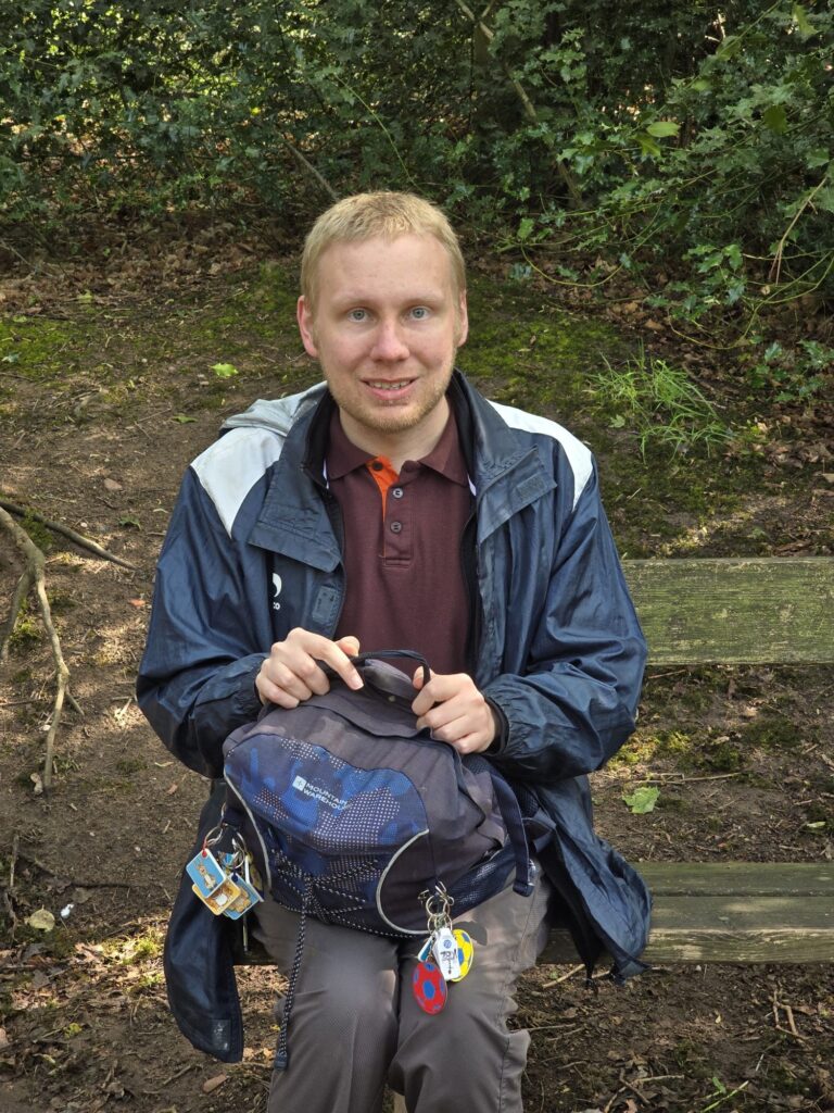 A man smiling and holding his rucksack while seated on a bench surrounded by trees.