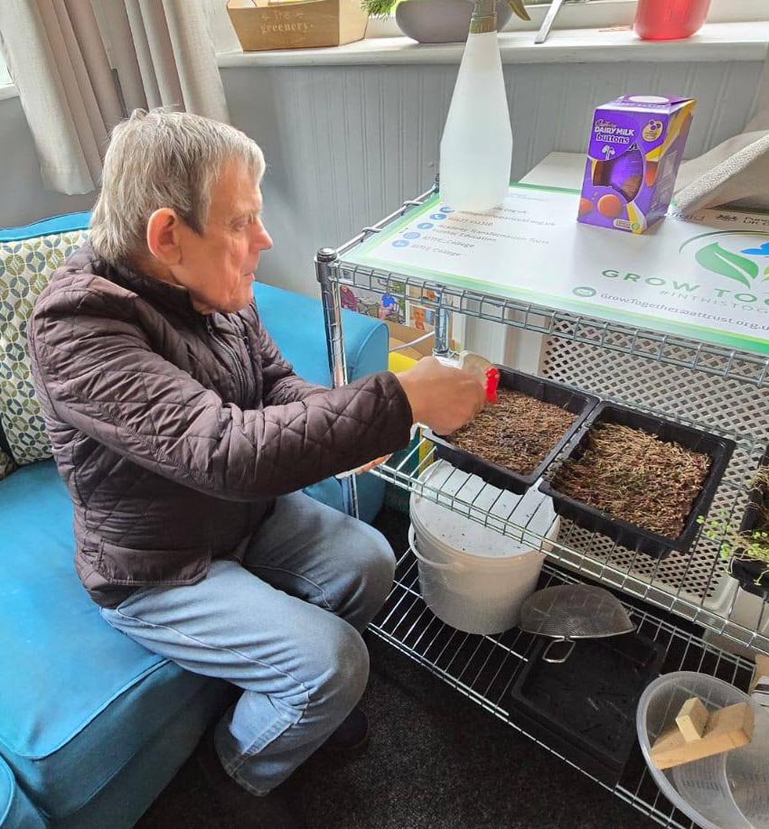 Watering seedlings indoors – A man sprays a tray of young seedlings using a red spray bottle on a shelf unit indoors, with an Easter egg box visible nearby.