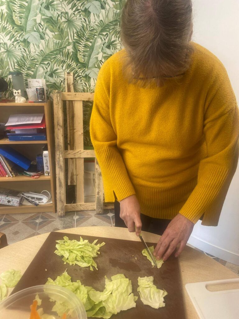 A woman in a yellow jumper finely slices cabbage on a chopping board at a table, concentrating on preparing ingredients for coleslaw.