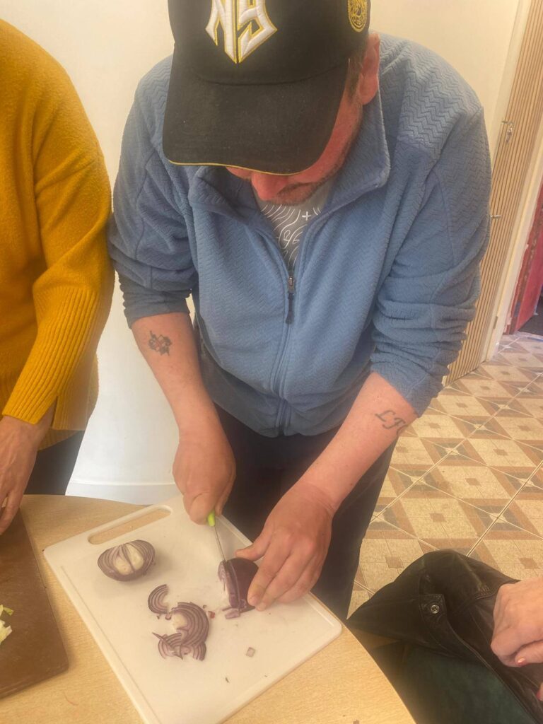 A man in a blue fleece and black cap carefully chops a red onion on a white chopping board during a group cooking session.