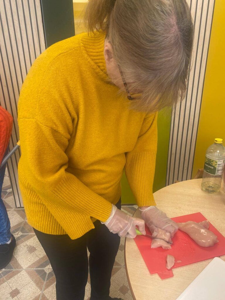 The same woman in a yellow jumper uses a knife to cut raw chicken breast on a red chopping board while wearing disposable gloves.