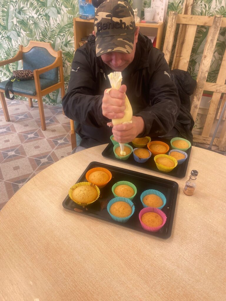 A man in a camouflage cap pipes icing onto freshly baked cupcakes in colourful cases, smiling as he decorates them.