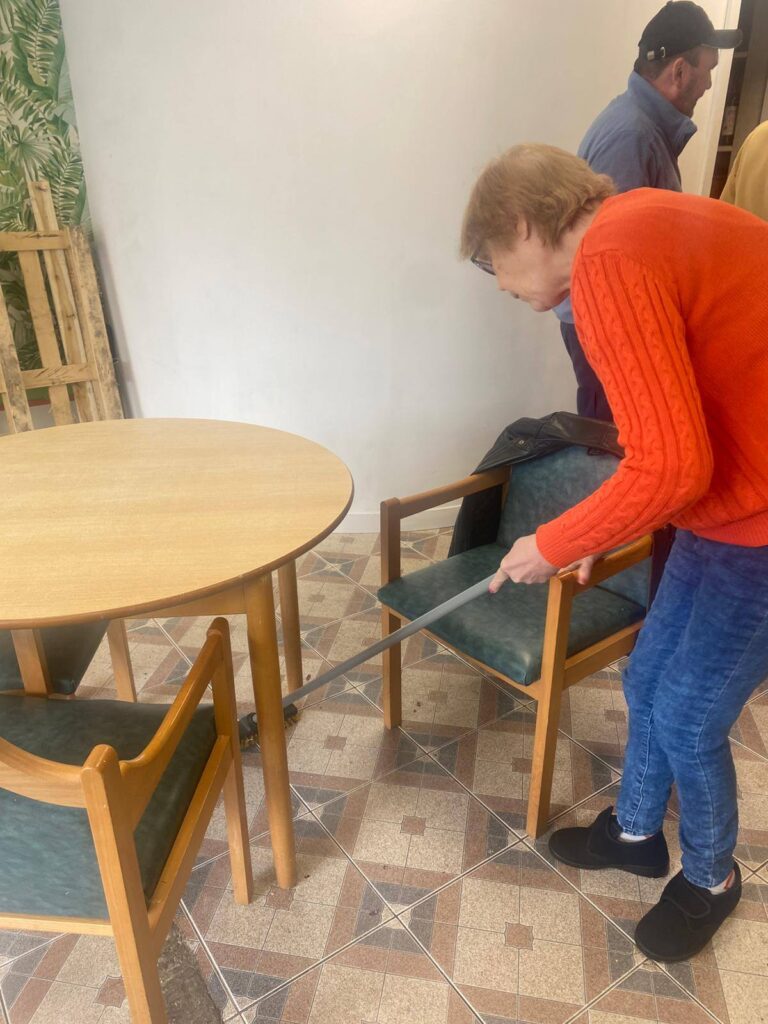 A woman in an orange jumper sweeps beneath a table with a long-handled dustpan brush, helping tidy up after cooking.