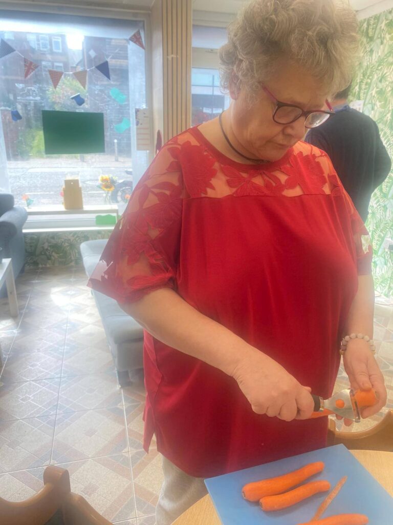 The same woman in red peels fresh carrots with a vegetable peeler beside a window decorated with colourful bunting.