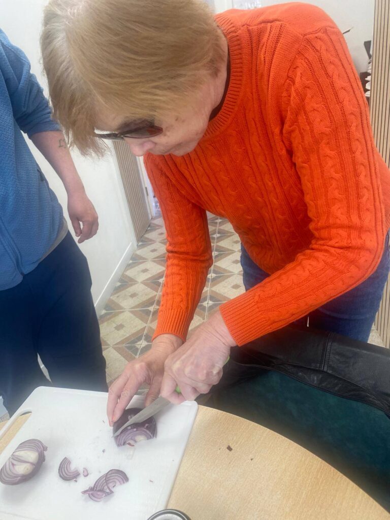 A woman in orange concentrates as she slices red onion on a white chopping board while another participant looks on supportively.