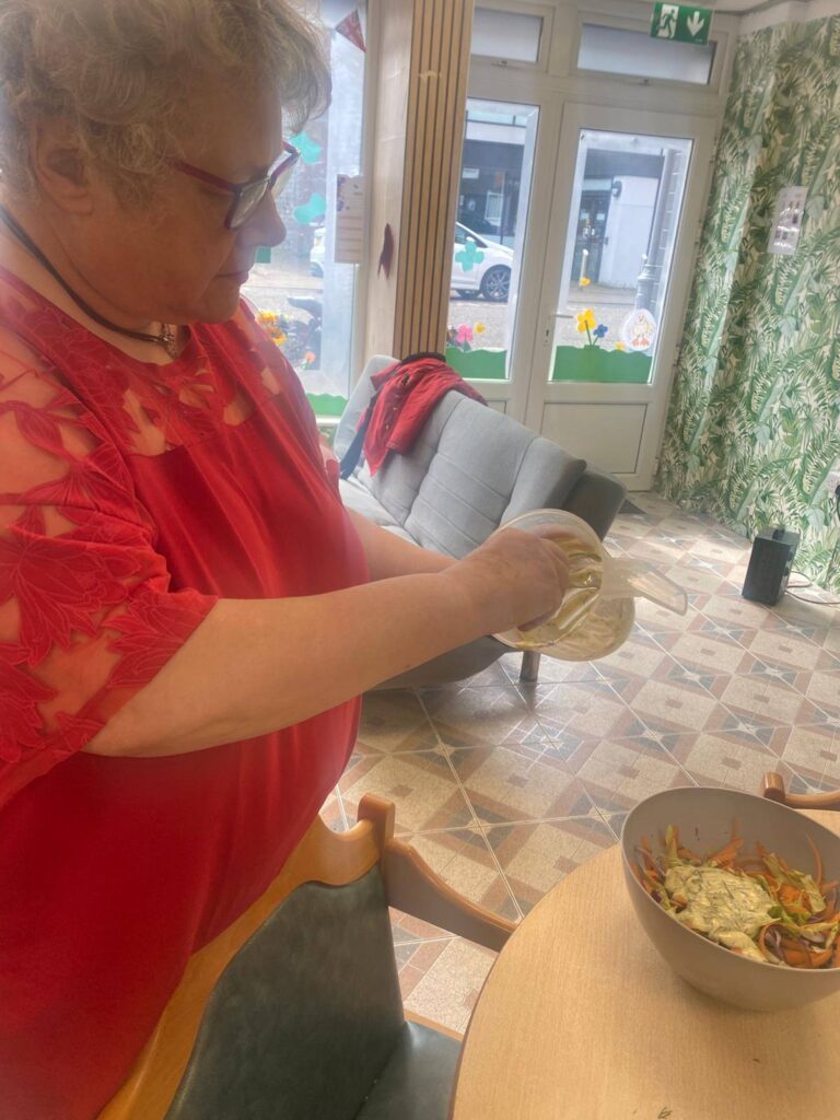The woman in red pours creamy dressing over a bowl of shredded vegetables to finish off a homemade coleslaw in a bright kitchen space.