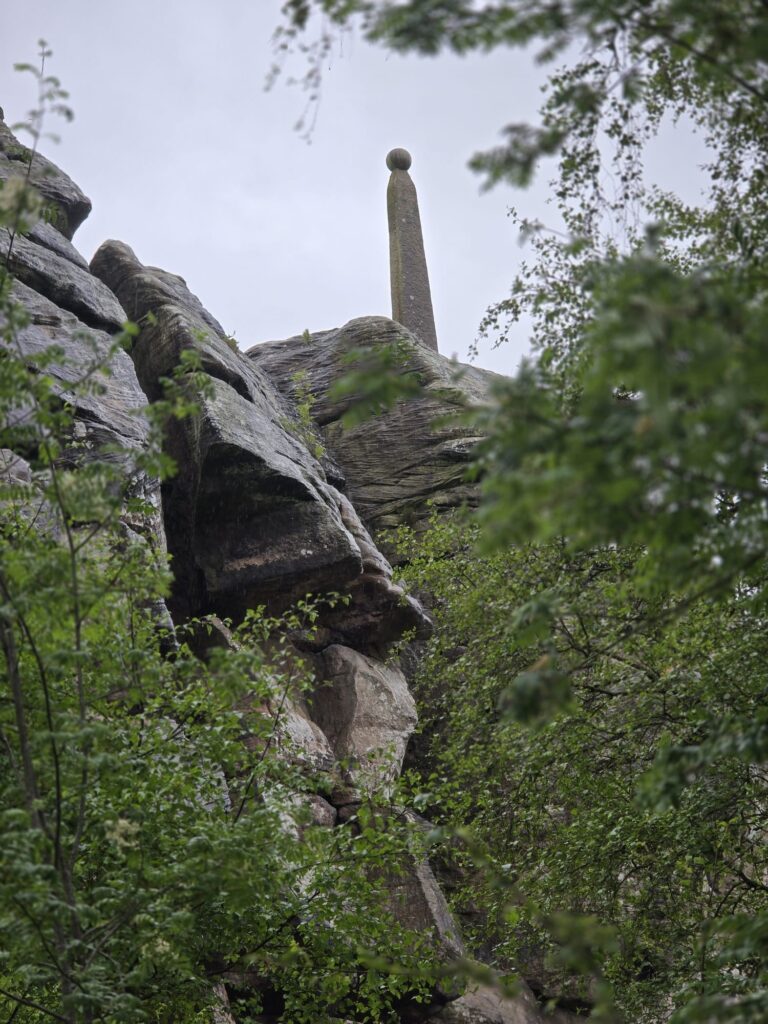 The Lord Nelson Monument seen through leafy green branches, partially obscured by surrounding rocks.