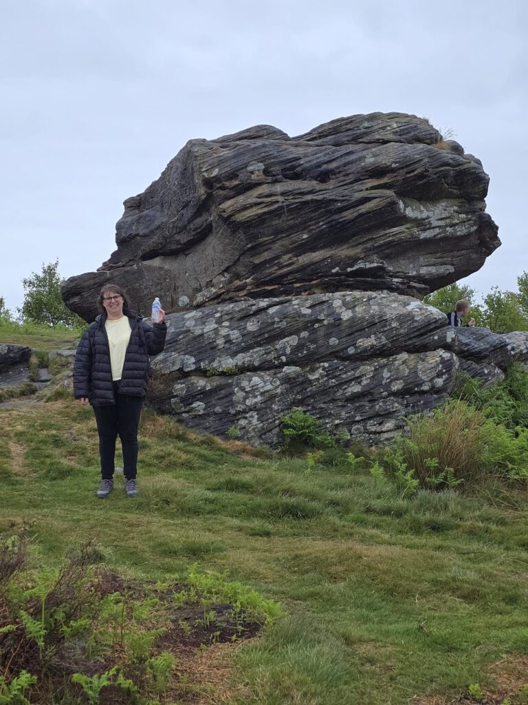 A woman stands smiling beside a large weathered rock formation, holding a water bottle, on grassy terrain.