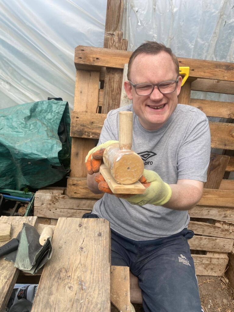 A smiling man in gloves holds up a handmade wooden steam train project while sitting at a rustic wooden workbench.
