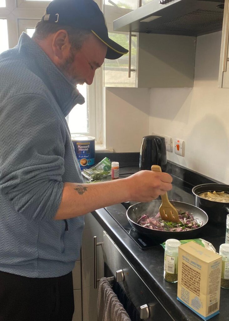 A man wearing a blue fleece and black cap stirs onions and greens in a frying pan on the hob. Ingredients including herbs and seasoning are visible on the worktop beside him
