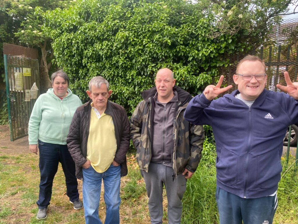 A group of four Gardening Club participants stand smiling in front of green hedging near the allotment gate.