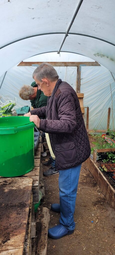 A man inside a polytunnel stands at a table filled with soil and trays, concentrating on a planting task.