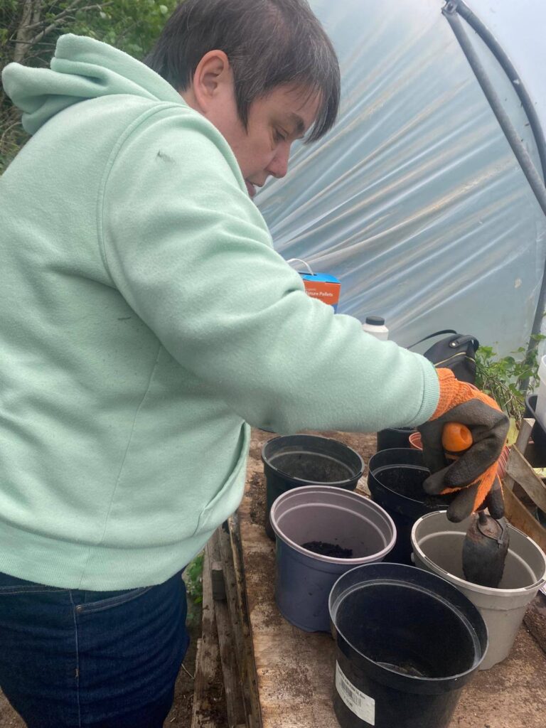 A woman in a mint green hoodie repots plants in a polytunnel, using gloved hands to handle small pots and compost.
