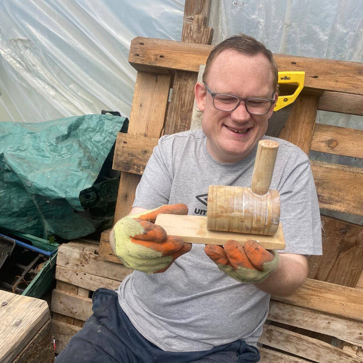 A close-up of a man proudly showing the wooden steam train he built, smiling at the camera with tools on the bench beside him.