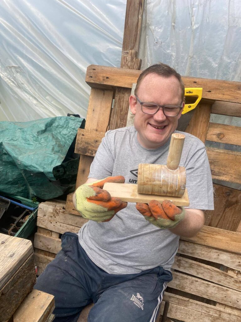 A close-up of a man proudly showing the wooden steam train he built, smiling at the camera with tools on the bench beside him.