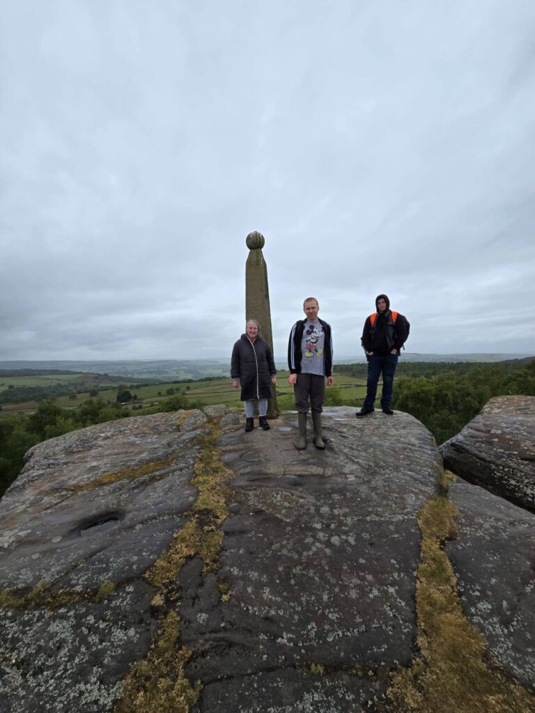 Three people stand on a large flat rock at the summit, with the Lord Nelson Monument and a panoramic countryside view behind them.